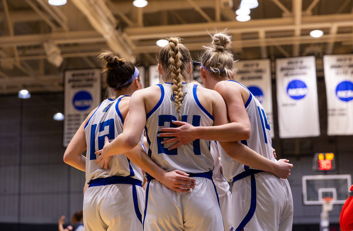 A group of basketball players in white and blue uniforms huddle together on a court, with their arms around each other.